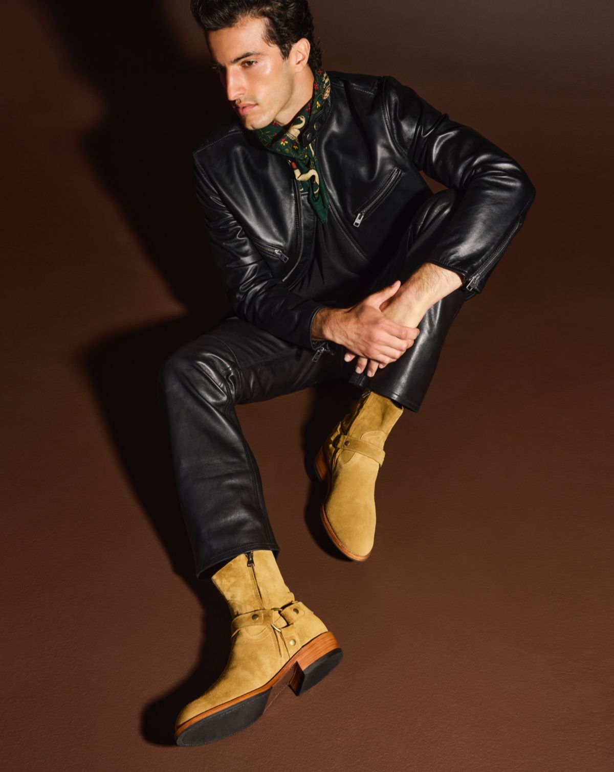 A man sitting down wearing a black leather jacket, black leather pants, and a pair of tan suede moto boots.  