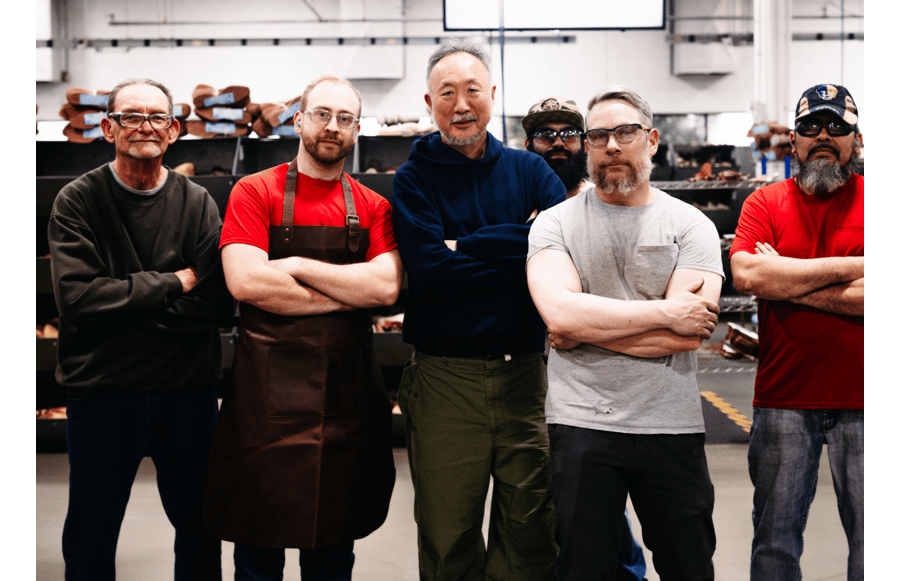 Daiki Suzuki and his team standing in a shoe workshop with arms crossed, with shelves of leather materials and tools in the background.