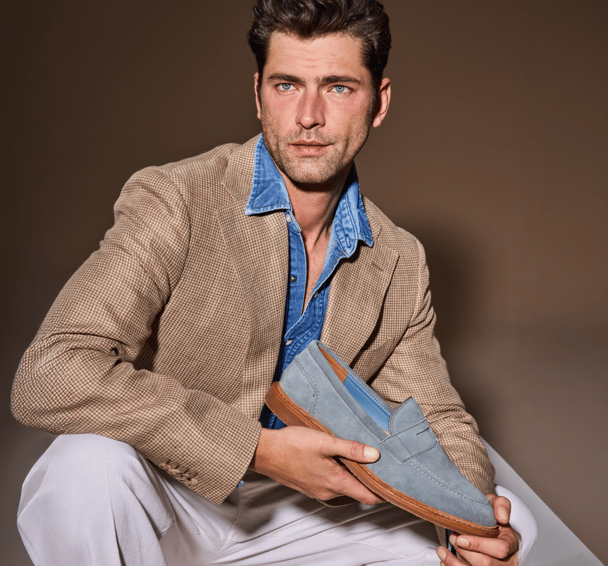 Man holding an Allen Edmonds Steel Blue Suede Randolph Verse Penny Loafer, styled with white trousers, a denim shirt, and a tan blazer 