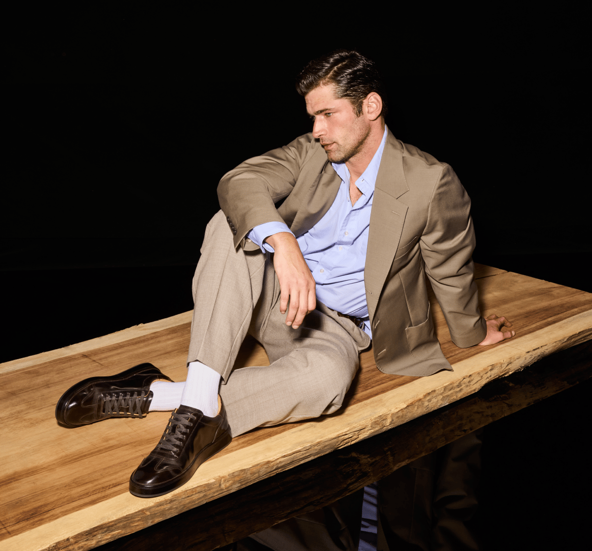 Dark brown Allen Edmonds Hawthorne sneakers and white socks worn by a man in a tan suit sitting on a wooden platform 