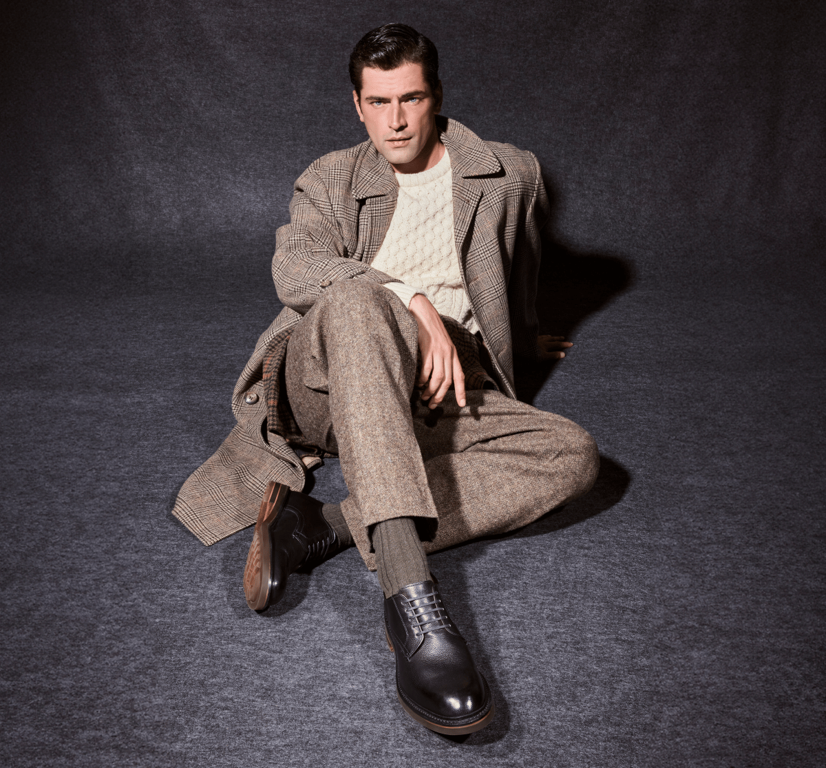 Black leather Allen Edmonds Noah Derby dress shoes and olive green socks worn by a man in a brown plaid overcoat and cream cable-knit sweater sitting on a grey background.