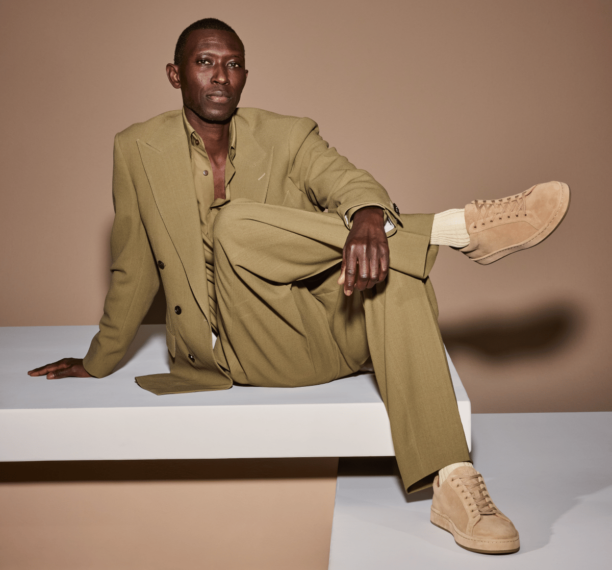 Tan suede Allen Edmonds Barnes lace-up sneakers and cream socks worn by a man in an olive green suit sitting on a white platform 