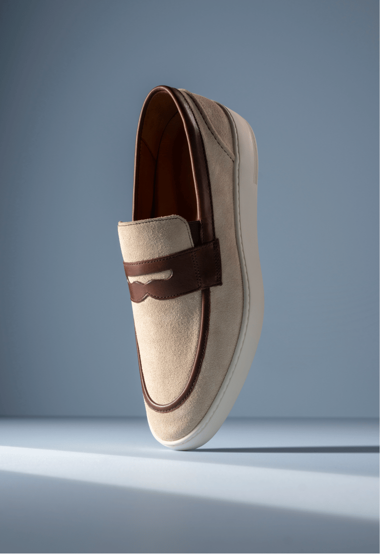 Tan suede Allen Edmonds Randolph 2.0 slip-on sneaker with brown leather trim and a white sole against a light blue background. 
